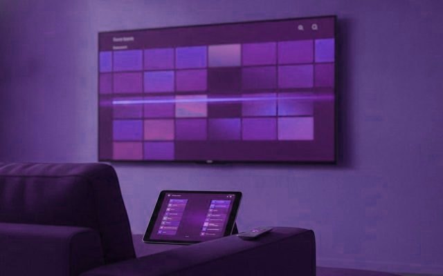 A TV showing a programme guide grid with a tablet and phone showing channel lists on a sofa