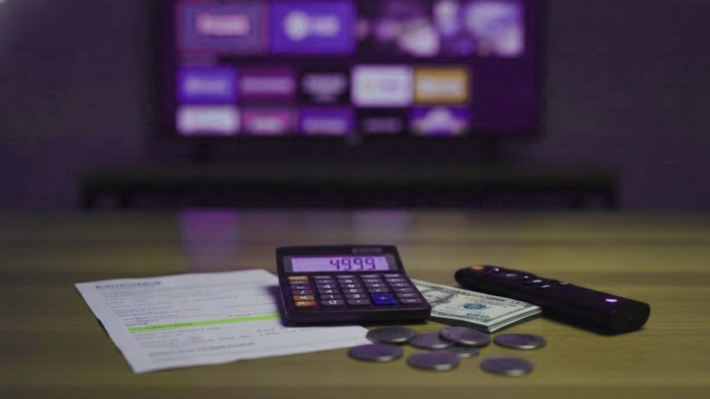 A calculator, dollar bills, coins, a streaming stick and a remote on a table in front of a TV